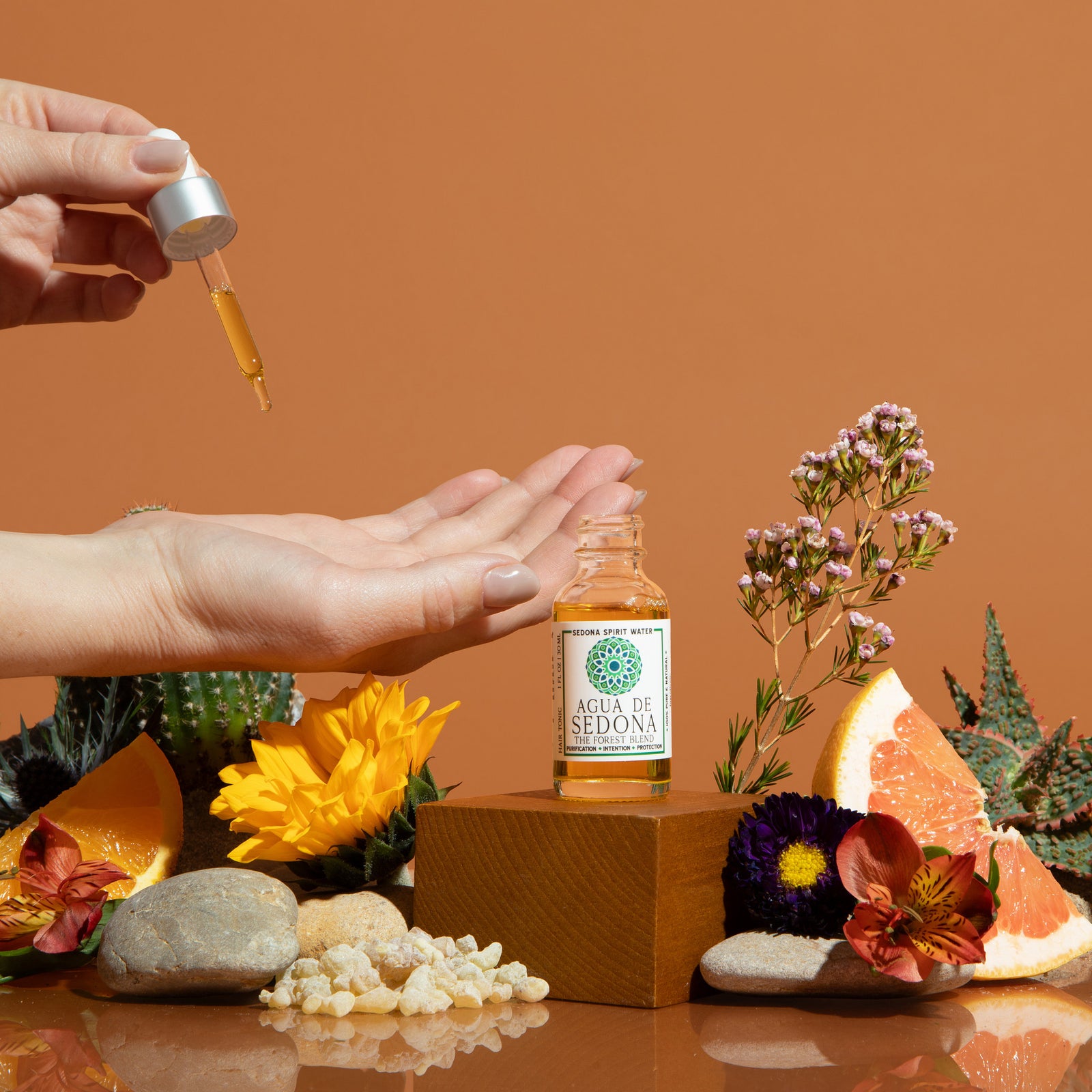 Agua de Sedona oil dropper in hand with citrus, flowers, and crystals on earthy orange backdrop