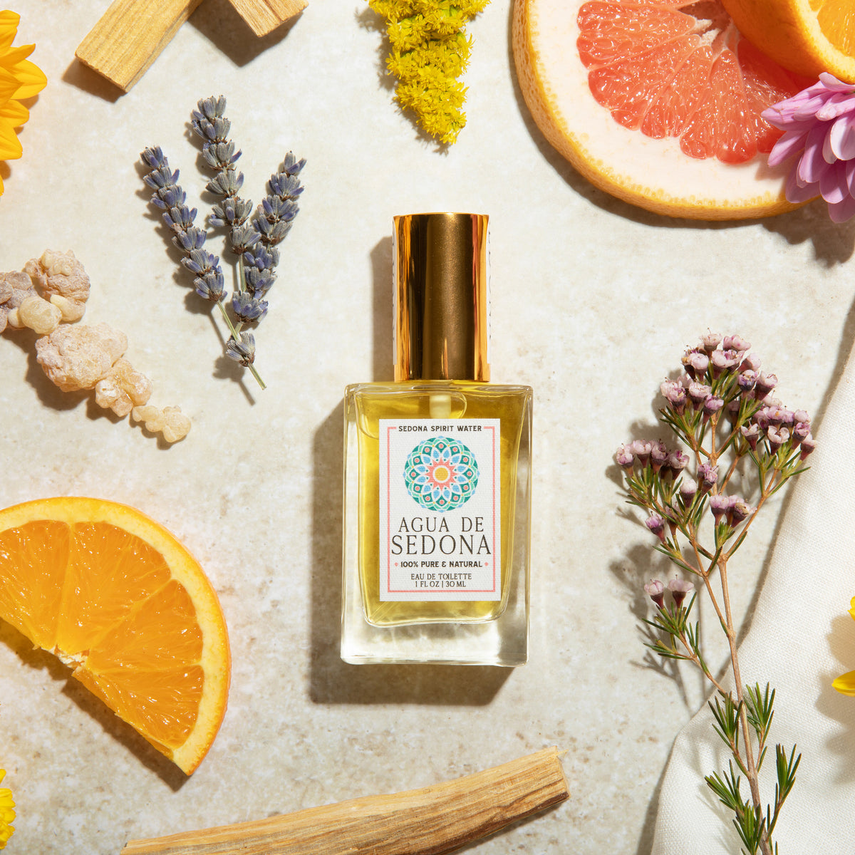 Agua de Sedona perfume bottle with gold cap, surrounded by citrus, florals, and natural elements.