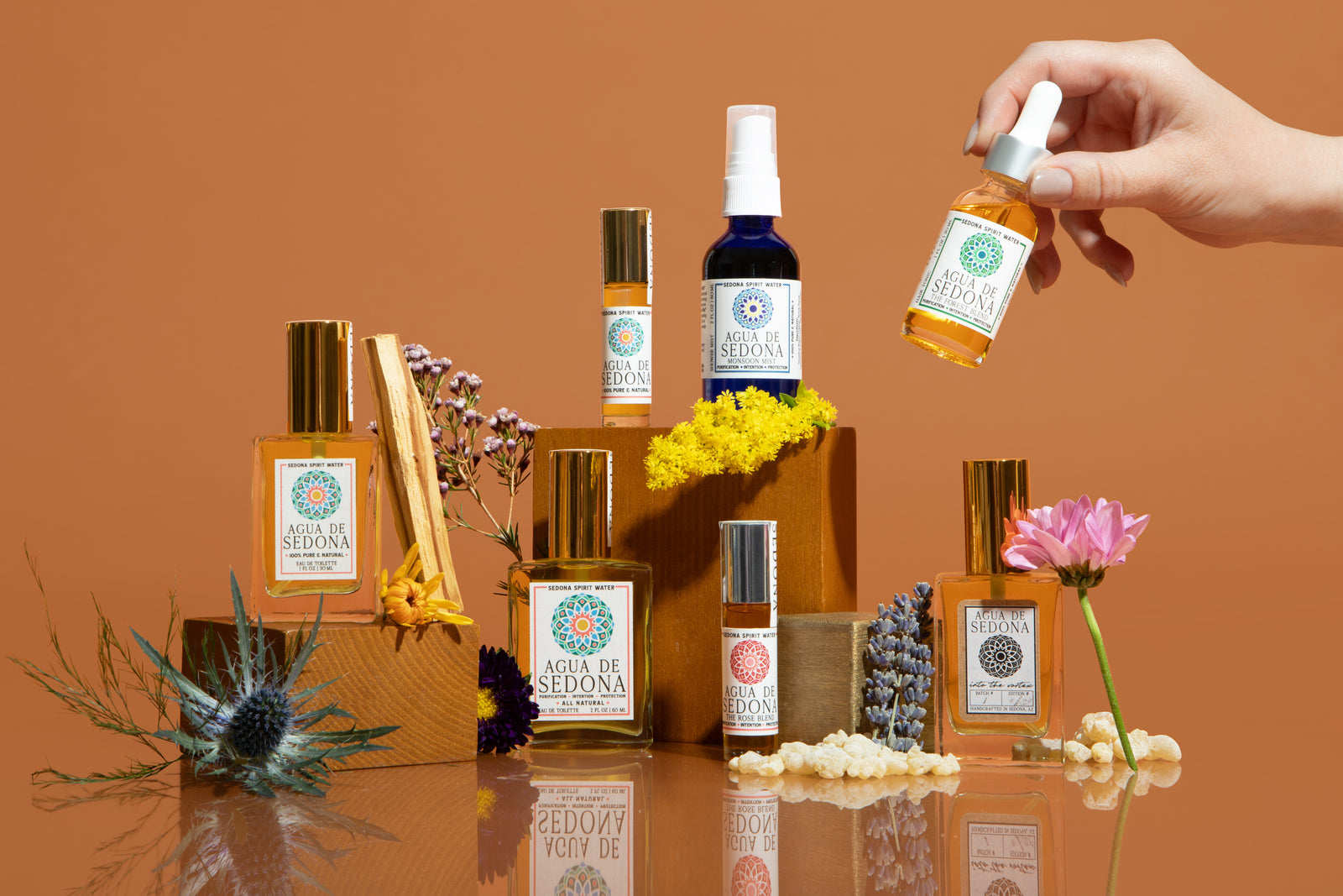 Collection of Agua de Sedona spirit water products with flowers, amber background, and hand holding dropper.