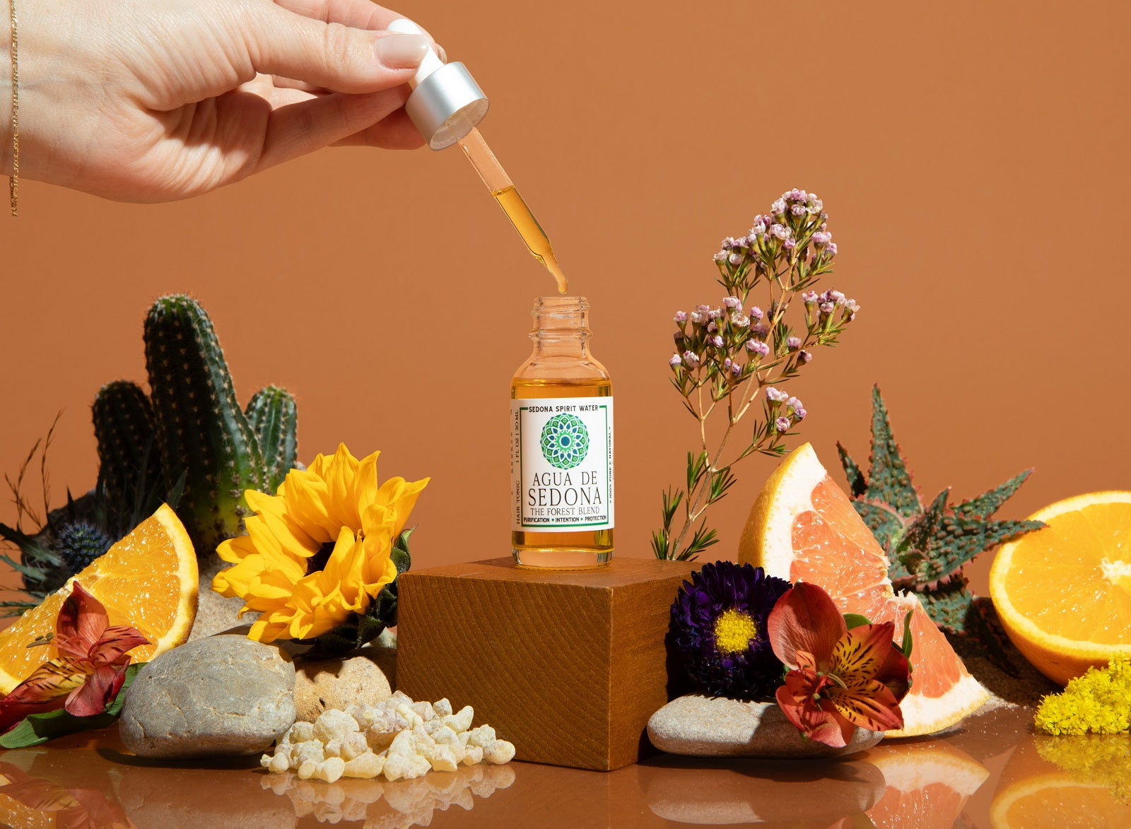Agua de Sedona - forest blend oil dropper in hand with citrus, flowers, and crystals on earthy orange backdrop