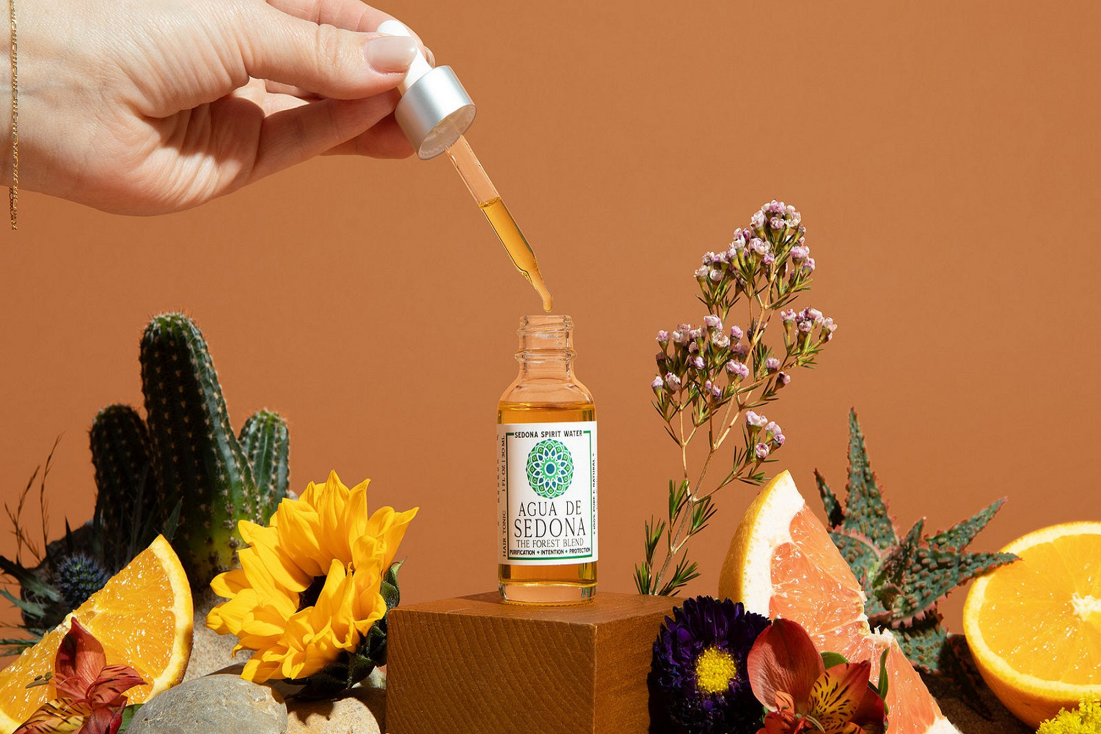 Agua de Sedona - forest blend oil dropper in hand with citrus, flowers, and crystals on earthy orange backdrop